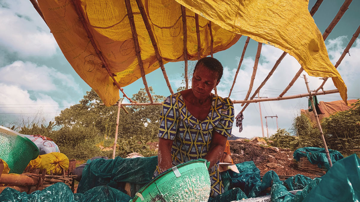 A person strains a mixture in a green colander under a yellow tarp in an outdoor setting.